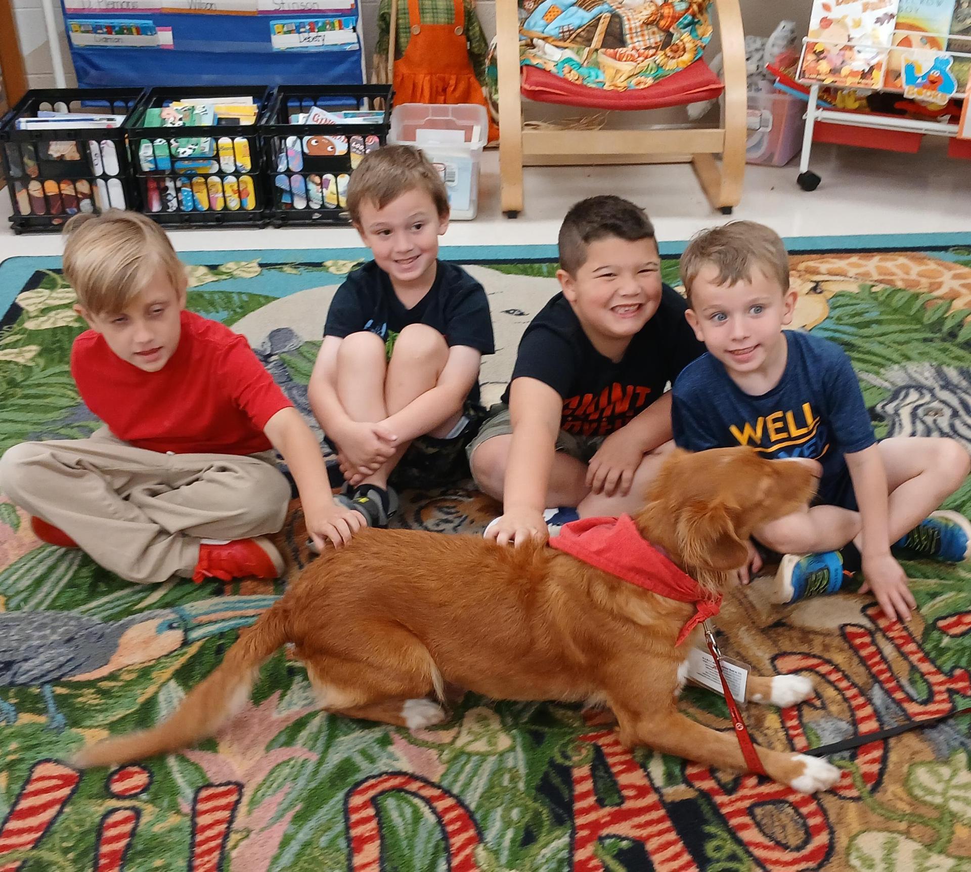 Four kids smiling while a dog moves across them, all seated on a vibrant classroom rug.