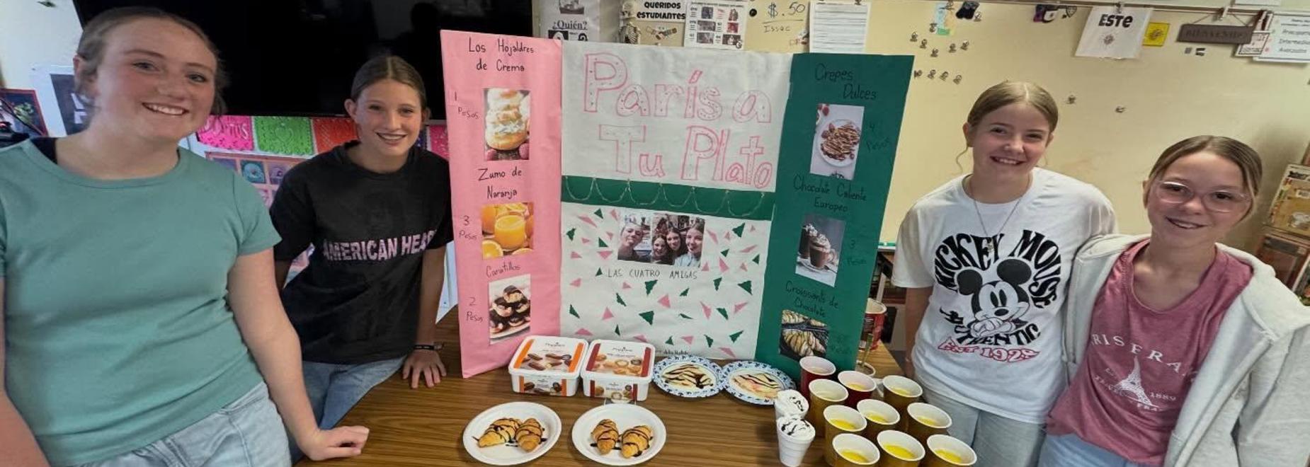Four girls gather around a project board displaying various treats and drinks.