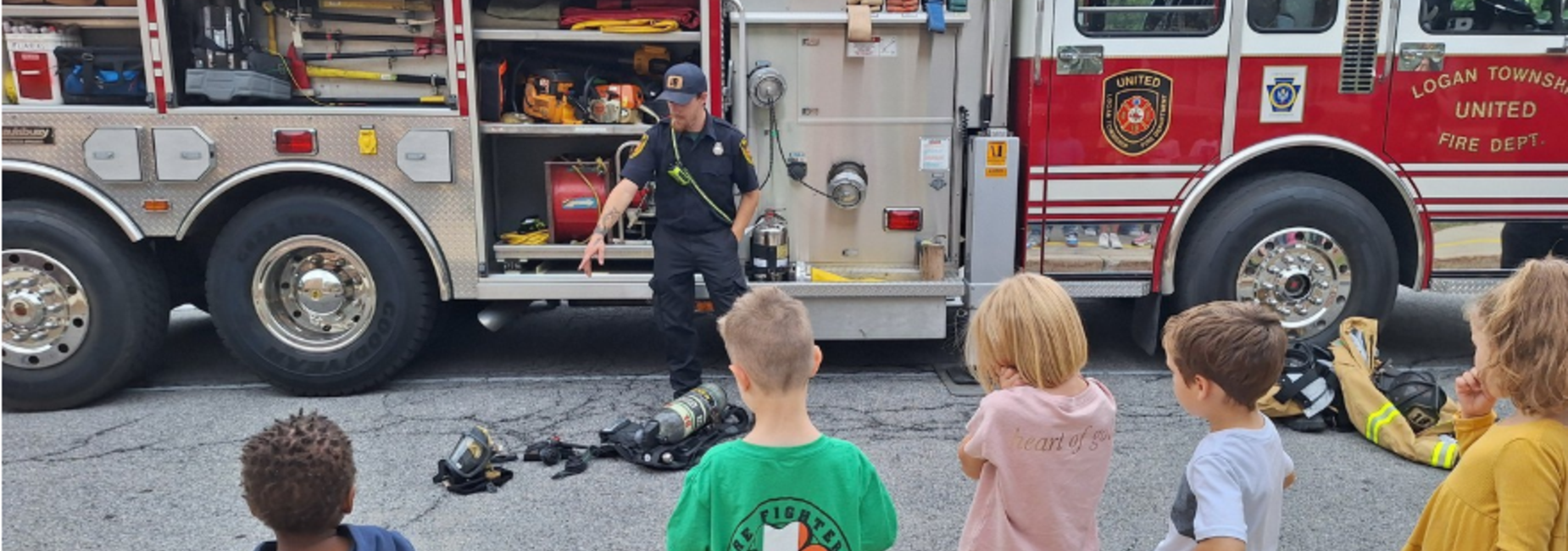 A firefighter instructing children near a fire truck and equipment on the ground.