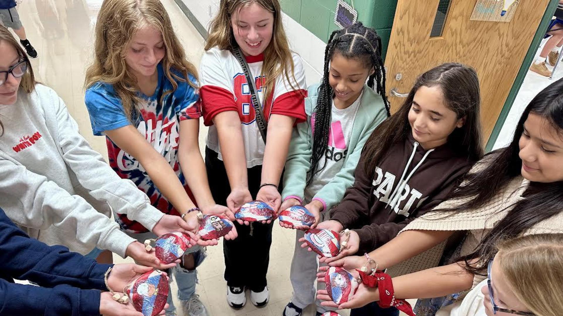 A group of diverse young girls holding painted rocks in a circle, smiling together.