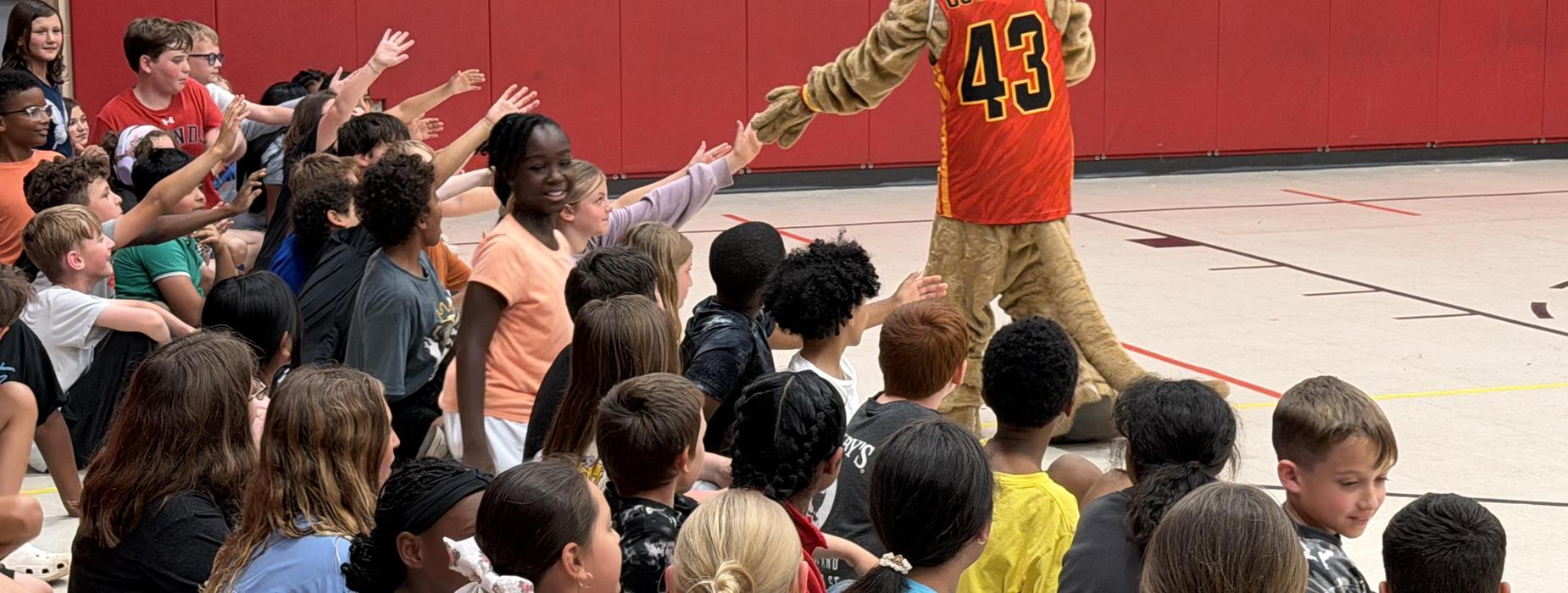 A large crowd of children and a mascot interacting in a gymnasium.