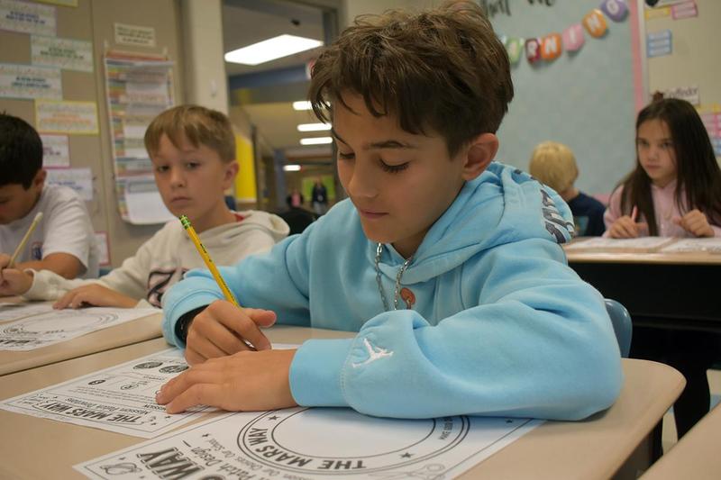 Boy writing at a desk, with another child and classmates nearby.