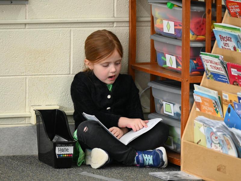Girl sitting on the floor reading a book beside colorful storage bins.