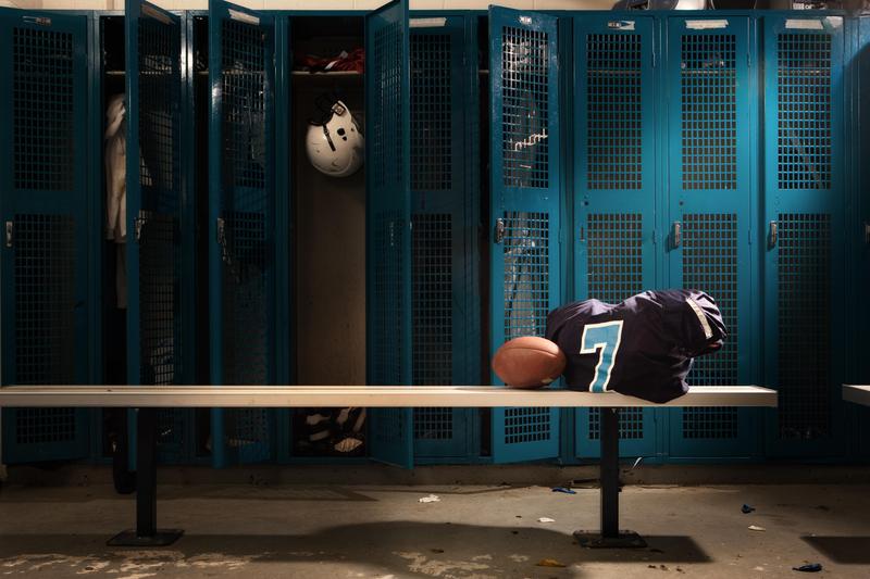 An empty sports locker room featuring a row of tall, teal mesh lockers. In the foreground, a long metal bench holds an American football propped against a folded navy blue jersey with a light blue and white number 7. One locker door in the background is open, revealing a white football helmet hanging inside.