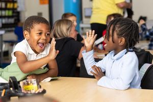 Two kindergarten students wave at each other and smile while seated at a table