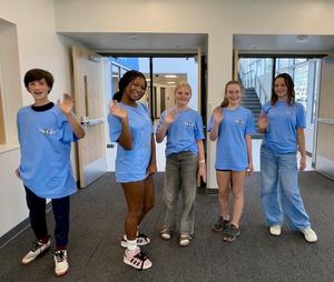 Five students post in blue t-shirts and wave to the camera