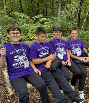 Four boys in matching purple shirts sitting on a swing in a wooded area.