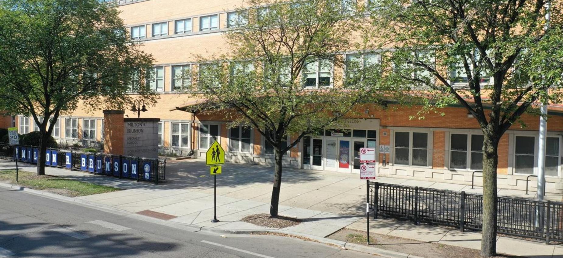 School building with trees and sidewalk in front, featuring signage.