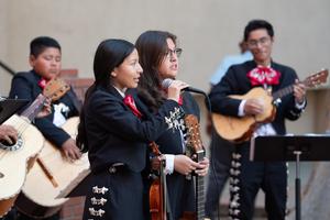 Two female student mariachi performers sing into a microphone while holding instruments, with other mariachi musicians playing guitars in the background.