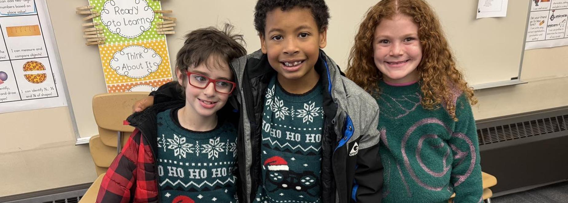 Three children in festive sweaters smiling together in a classroom setting.