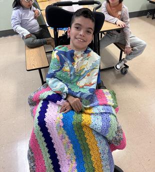 Boy in a wheelchair smiling, draped in a colorful blanket at a classroom.