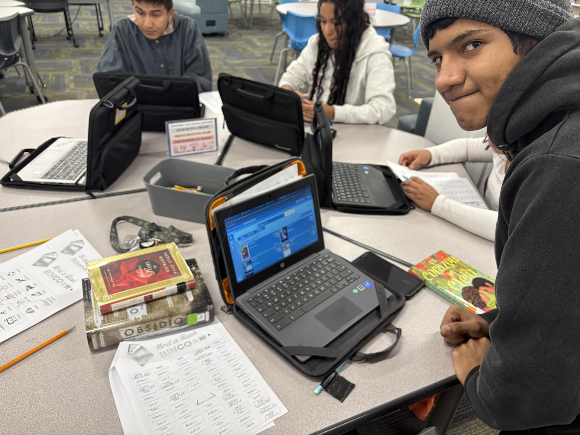 Students working on laptops at a table with books and homework materials.