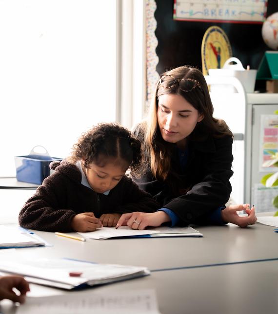 A child writes in a notebook while an adult guides her at a table.