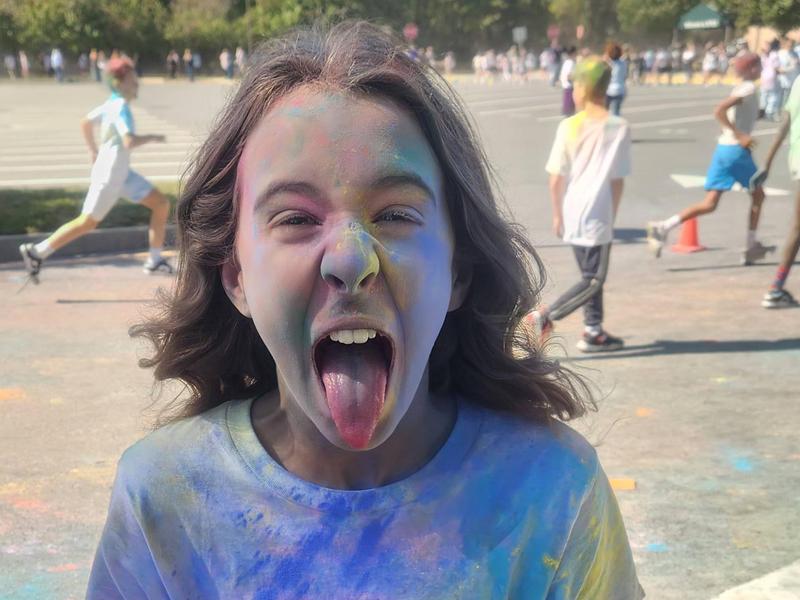 Child covered in colorful powder, sticking out their tongue, with a playful expression.