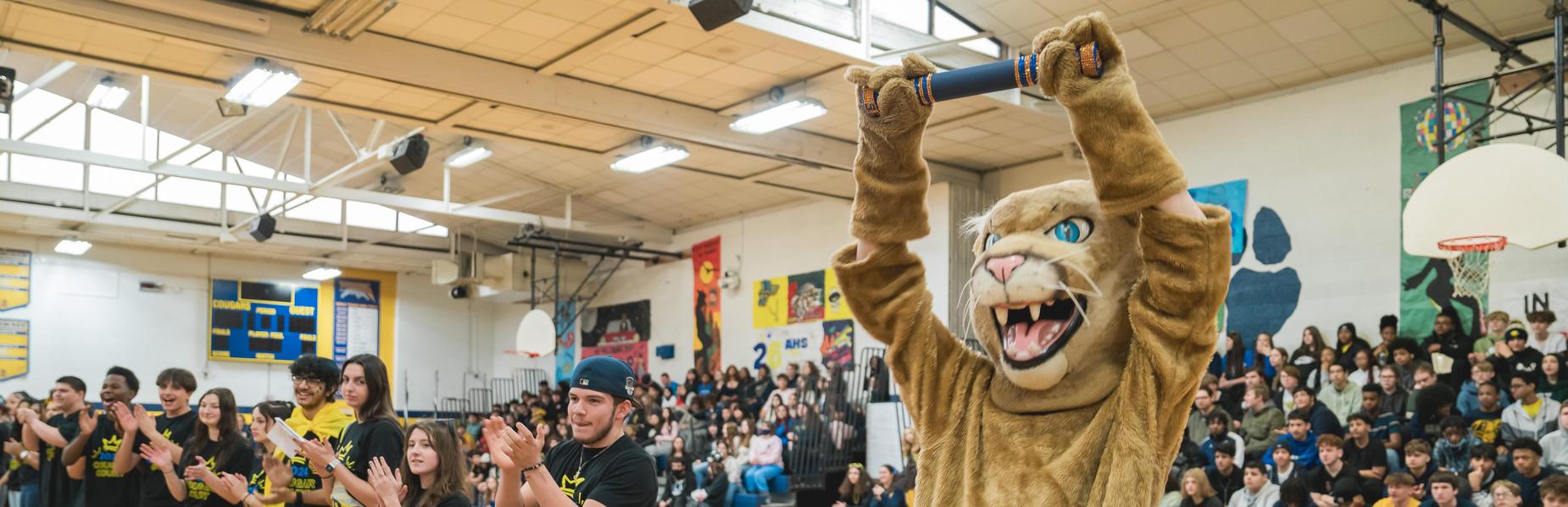 A school assembly with students cheering and a mascot on stage.