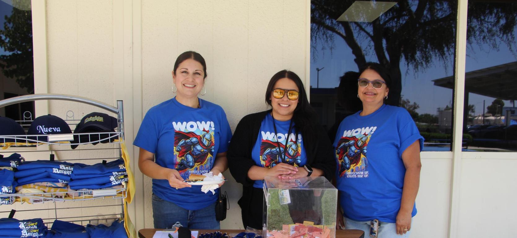 Three women wearing blue shirts with a table displaying raffle tickets and merchandise.