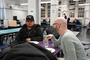 Two men sitting at a table, one drinking water and the other watching.