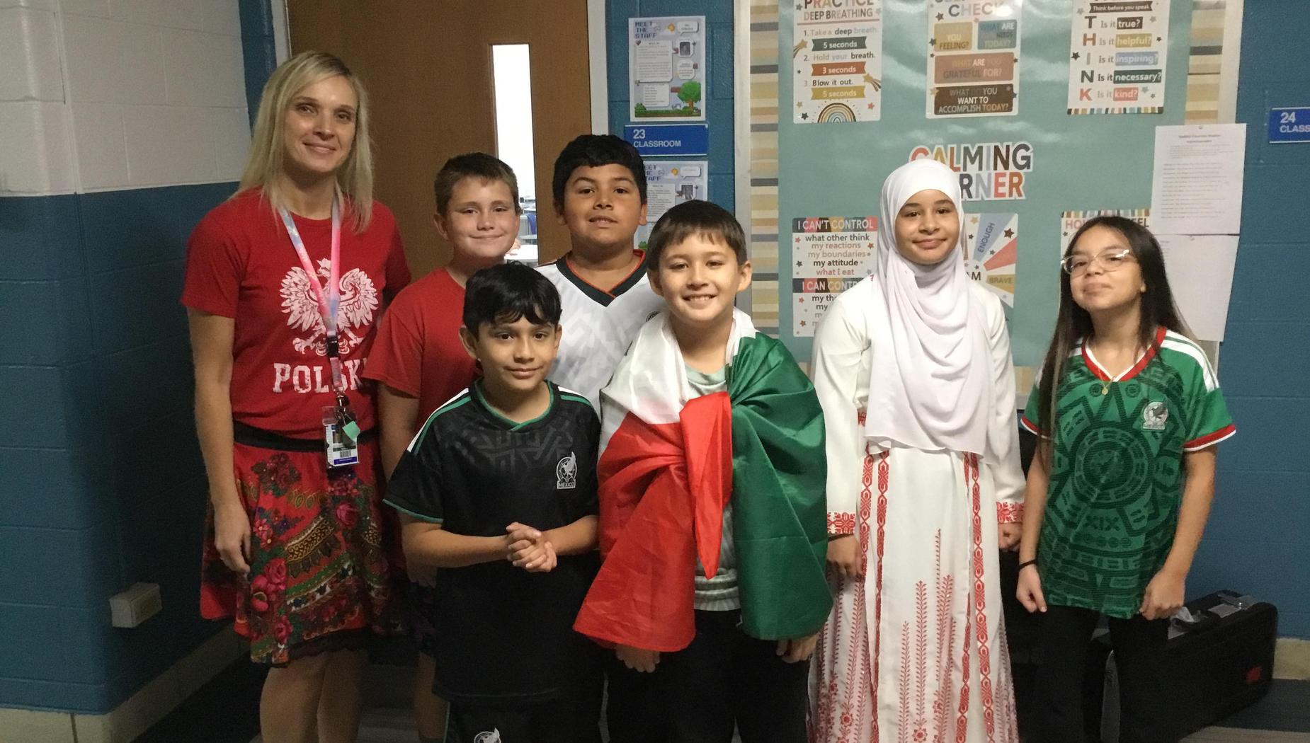 Children with flags and cultural clothing pose with a teacher in a decorated classroom.