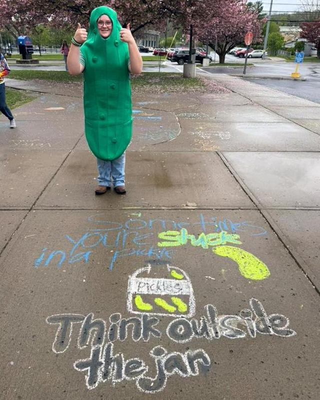 A student wearing a pickle costume gives a thumbs up next to a sidewalk chalk message.