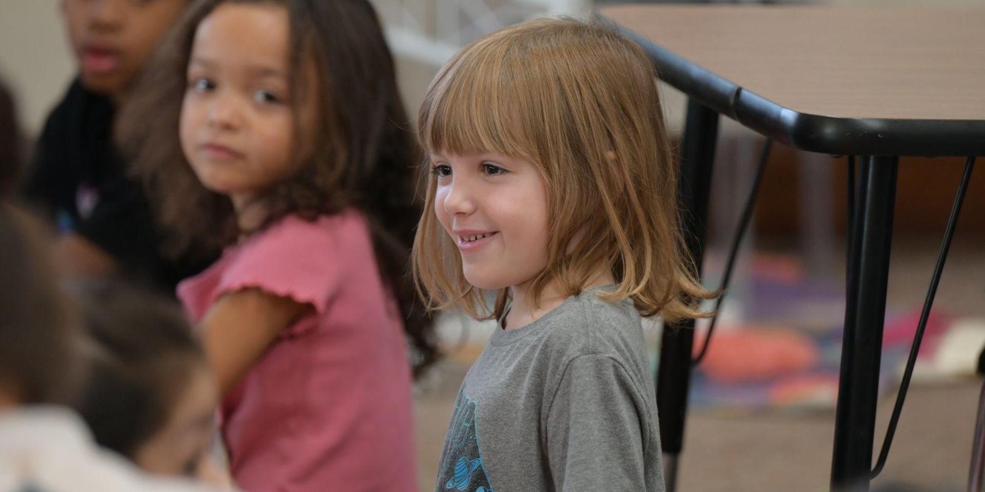 Smiling child in a classroom with peers in the background.