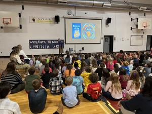 A speaker presents to a large group of children seated on the gym floor.