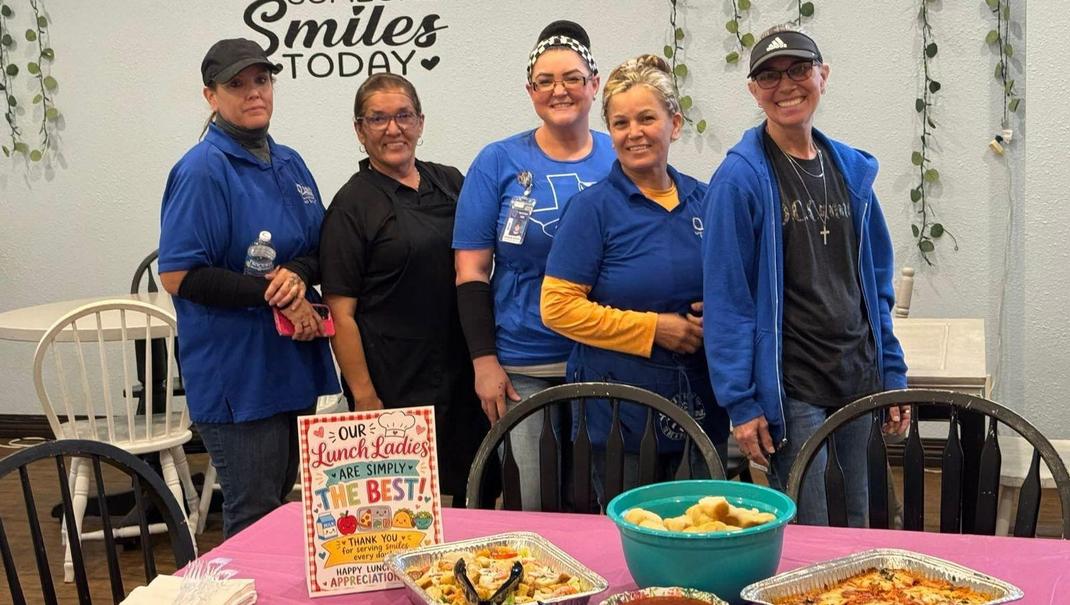 Five women in blue shirts standing by a table with food.