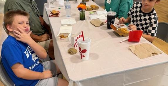 A table with two kids eating Chick-fil-A, a man in the background, and drinks.