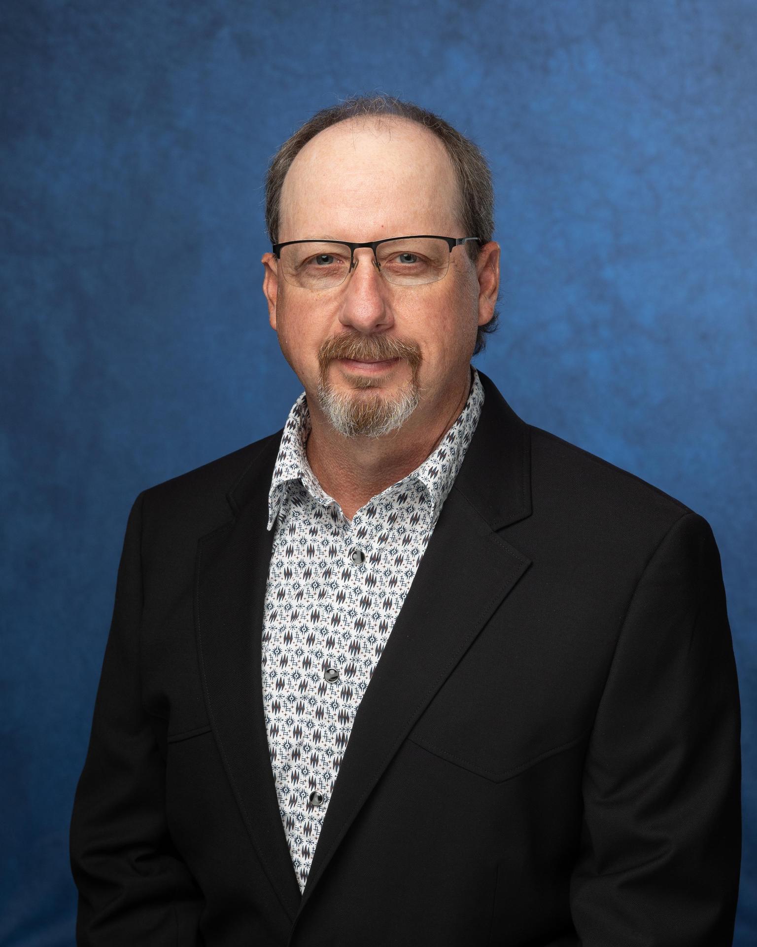Man with glasses, black suit and a patterned shirt in front of a blue background.