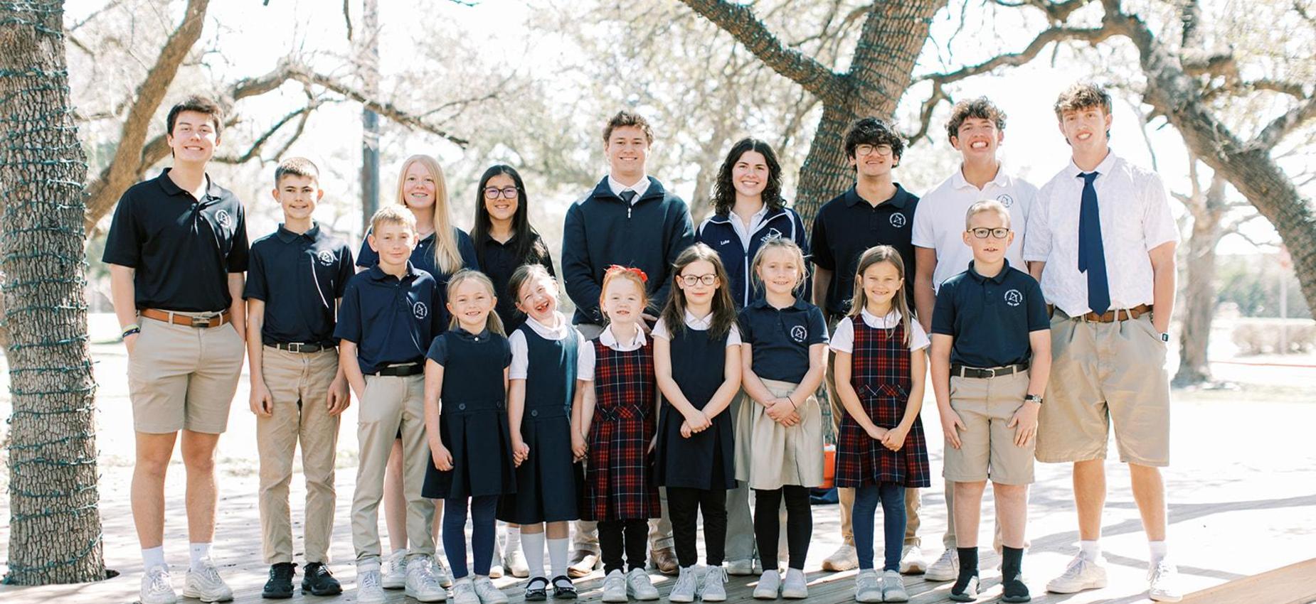Students in Founders uniforms, from the youngest grades through high school, stand together under oak trees