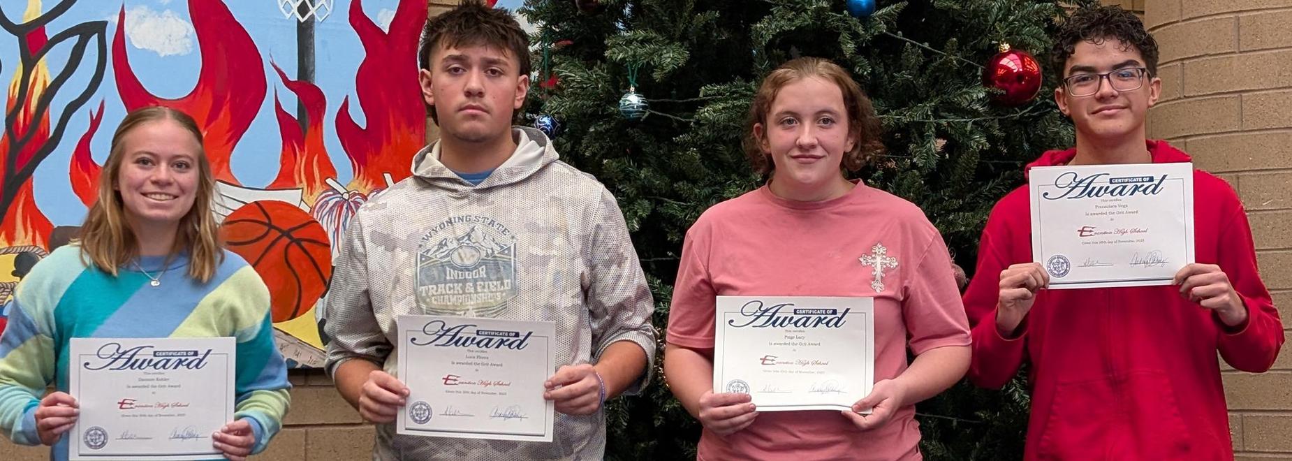 Four students holding award certificates in front of a Christmas tree.