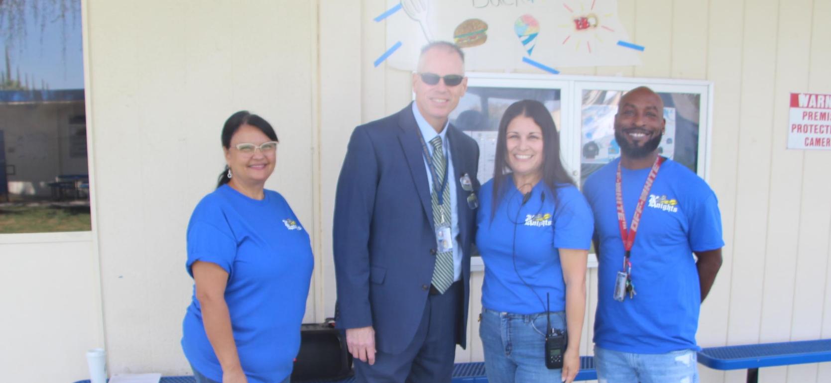 Four people standing together outside a building, smiling and wearing blue shirts.