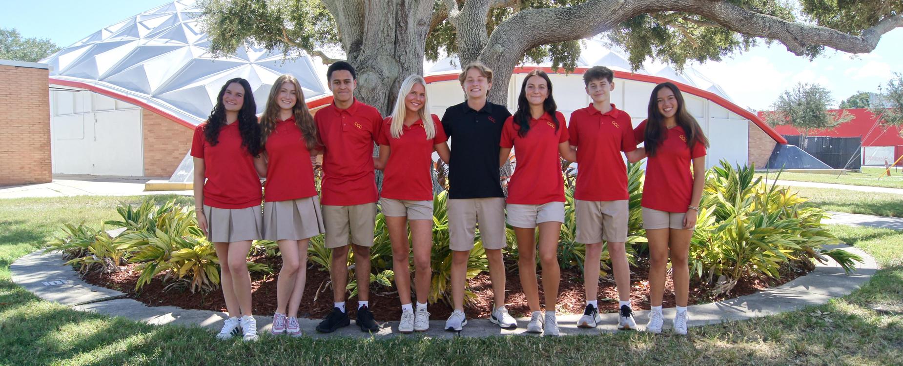 A group of students (five girls and three boys) stand together smiling arm in arm under a large tree in front of Cavallaro Gymnasium
