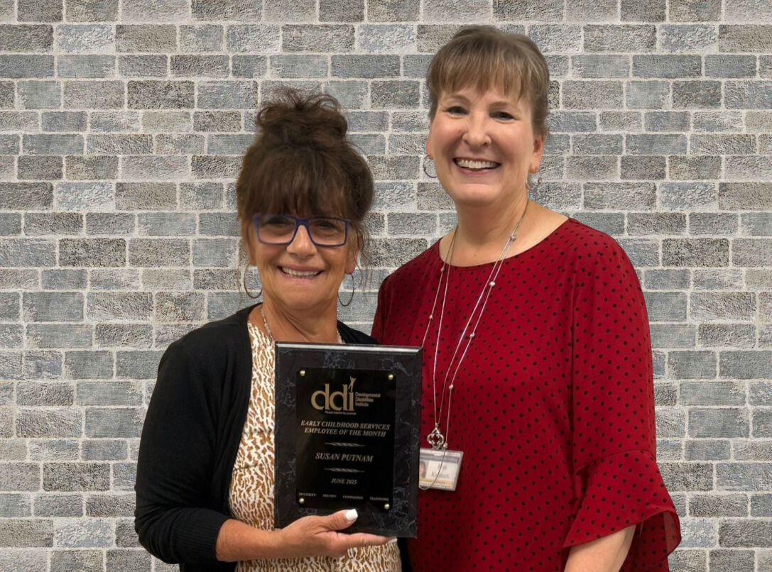 One woman standing with plaque smiling; one woman next to her smiling; blue/gray brick background