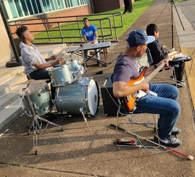 Students perform outdoors with drums, keyboard, and electric guitar on a sunny day.