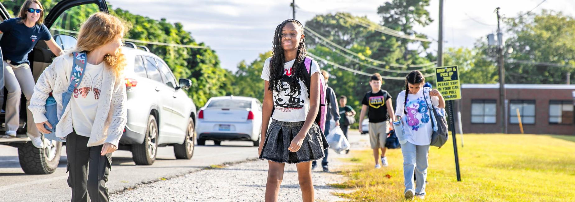 Students exiting vehicles and walking on the sidewalk towards school, smiling and chatting.