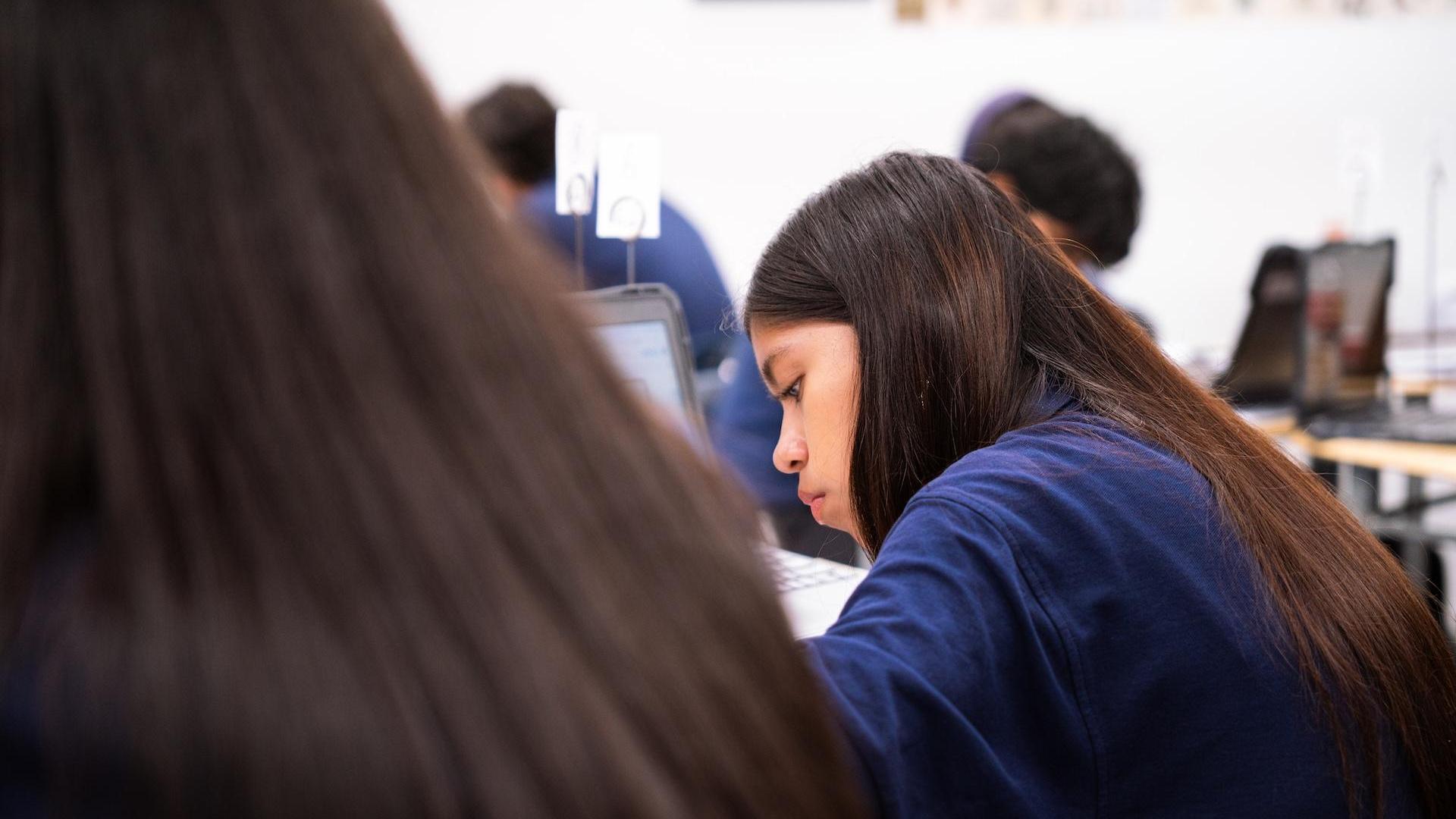 A student with long hair writing notes, focused in a classroom setting with peers in view.