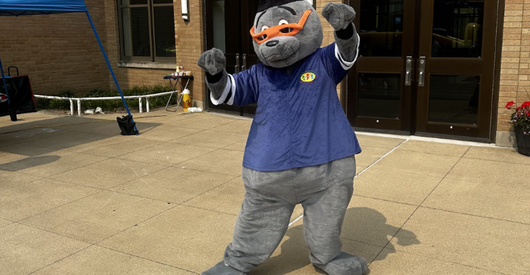 A gray cartoonish mascot wearing a blue shirt and orange glasses, dancing outdoors.
