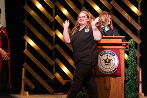 Graduate in black scrubs walks off stage with a joyful expression, raising her hands in celebration after receiving her certificate.