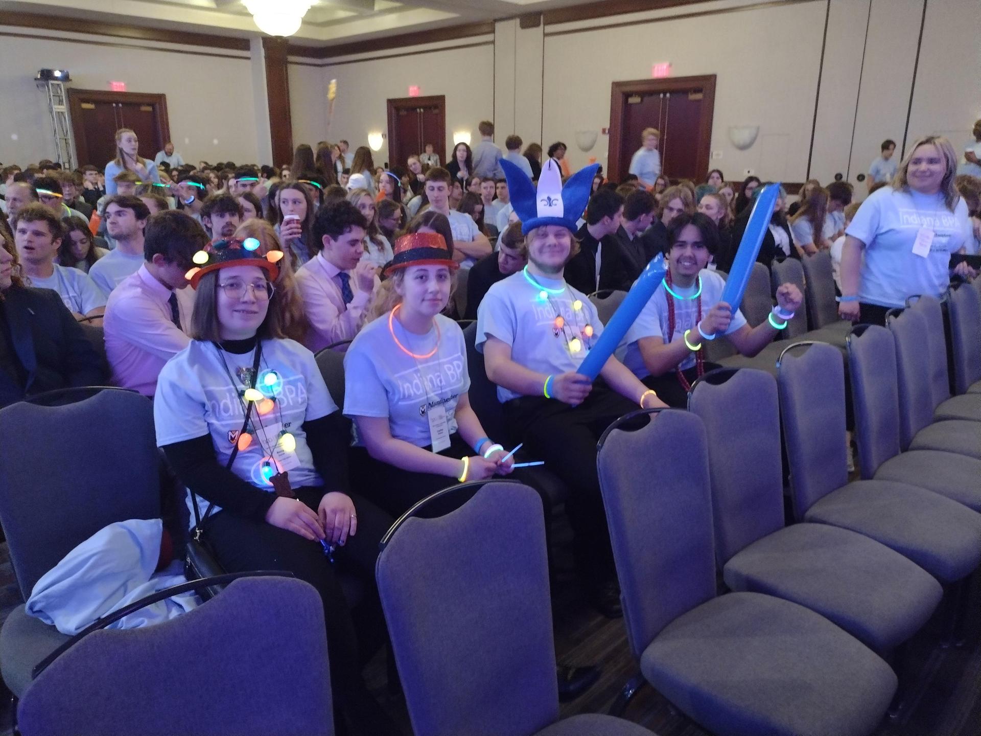 Students wearing glow accessories, sitting in a crowded conference hall.