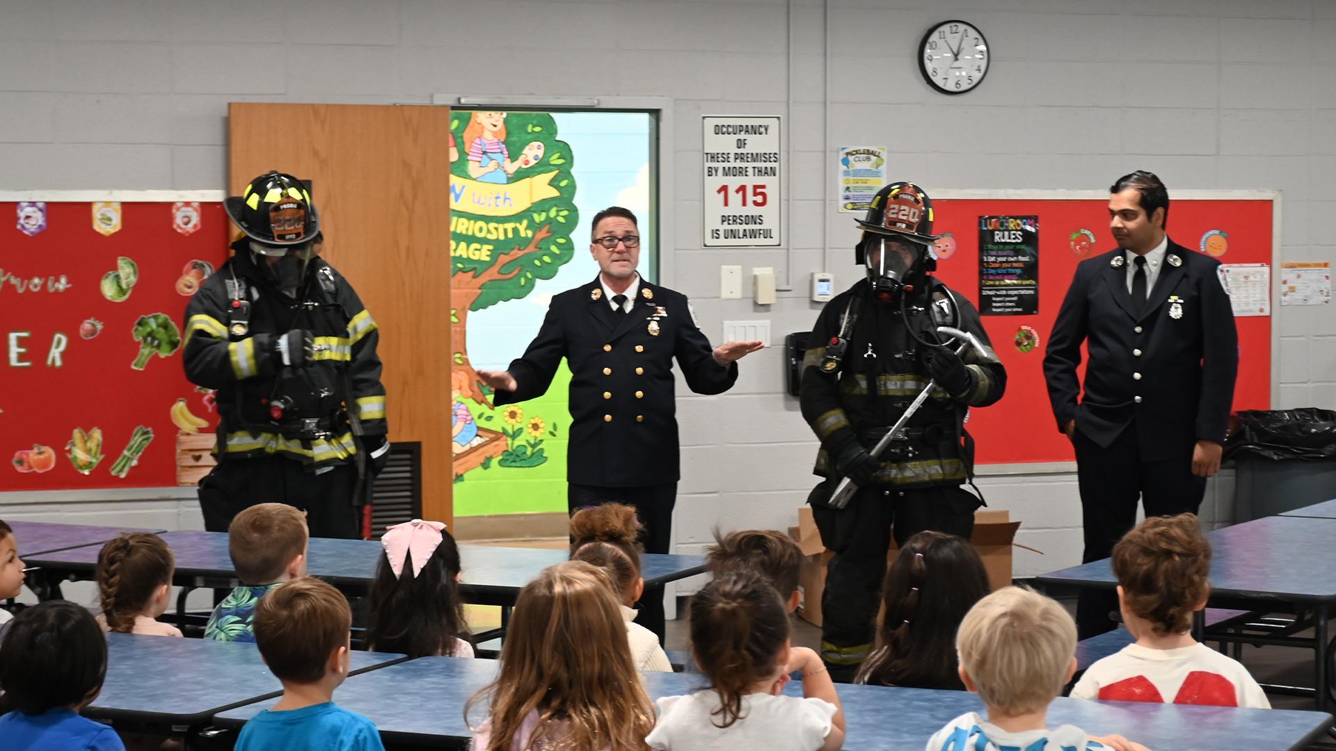 Firefighters speaking to children in a school cafeteria setting.