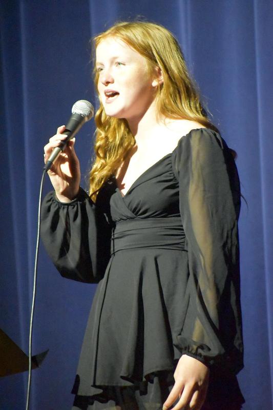 Girl with red hair performing into a microphone, dressed in black.