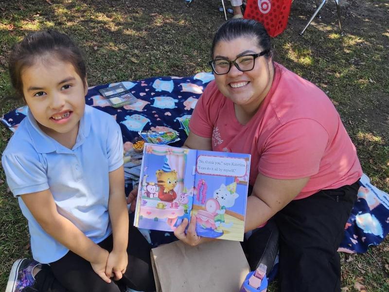 A woman and a girl sitting on a blanket, reading a children's book outdoors.