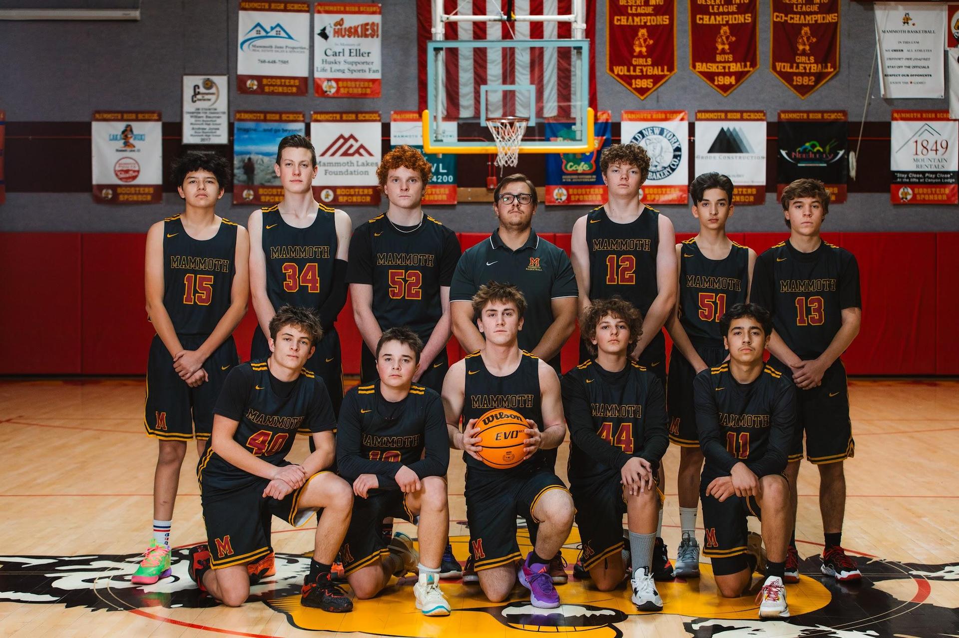 Basketball team in jerseys, posed on court with a hoop and banners behind them.