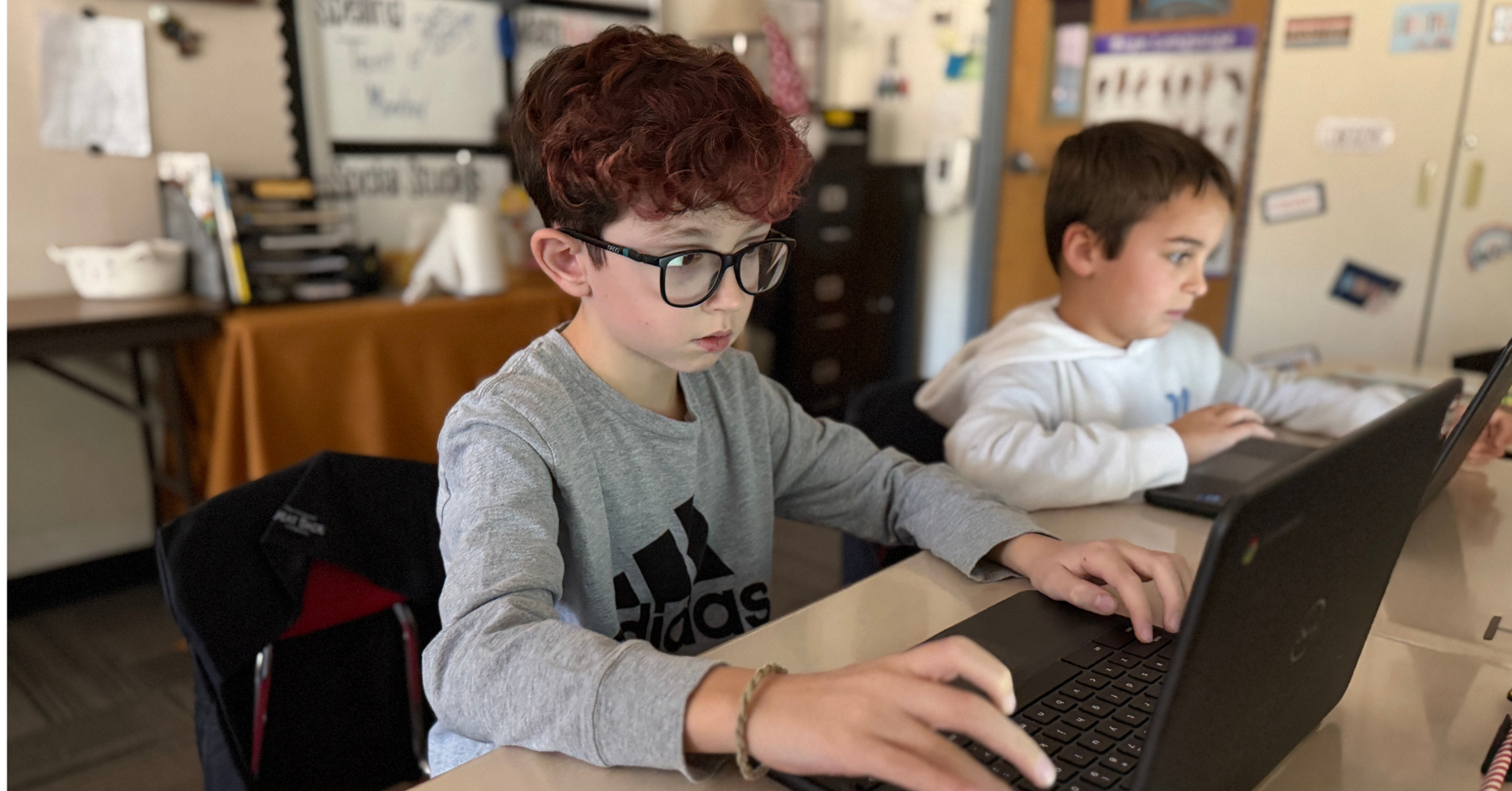 Two children working on laptops in a classroom setting.