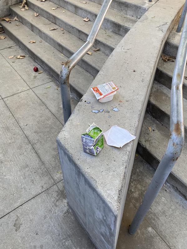 utdoor concrete staircase with metal handrails, littered with discarded food containers including an orange cereal cup and green milk carton, along with a crumpled napkin and dried leaves scattered across the steps and landing.