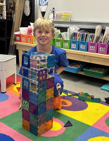 A smiling boy poses with a tall structure made from colorful building blocks on a play mat.