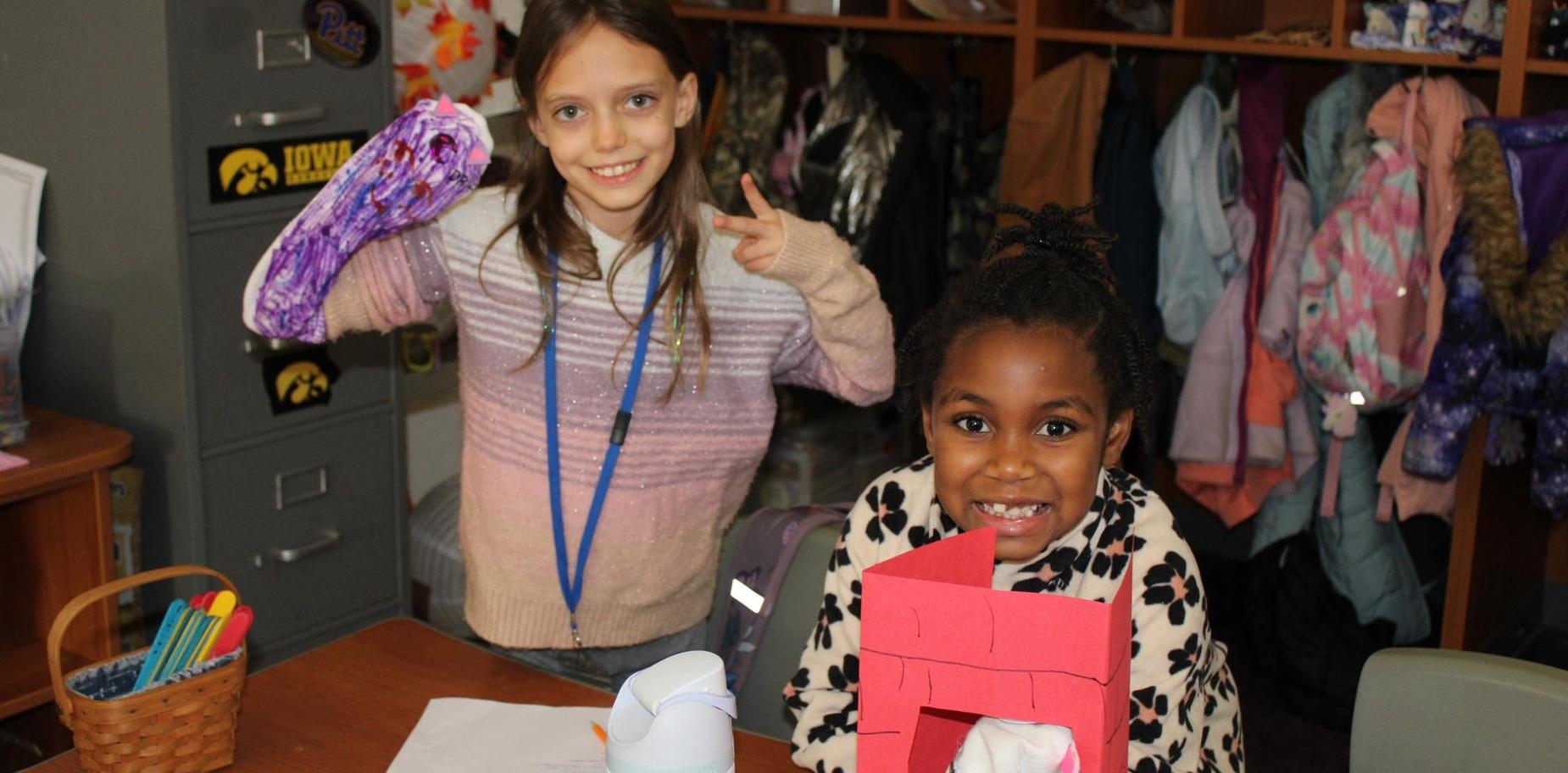 Two girls smiling while holding crafts in a classroom environment.