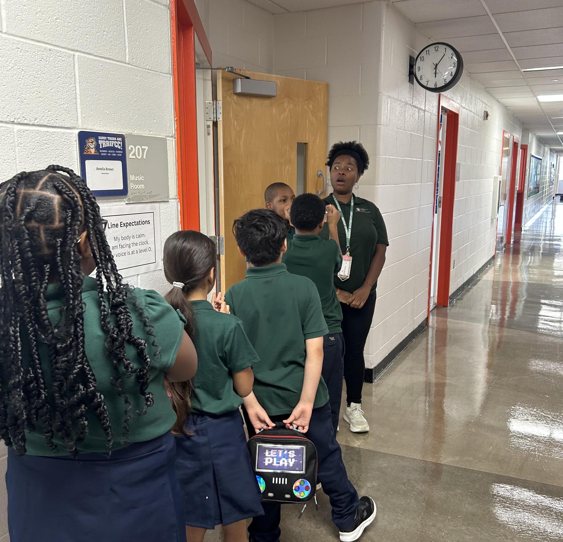 Students in green shirts line up outside a classroom, with one teacher guiding them.