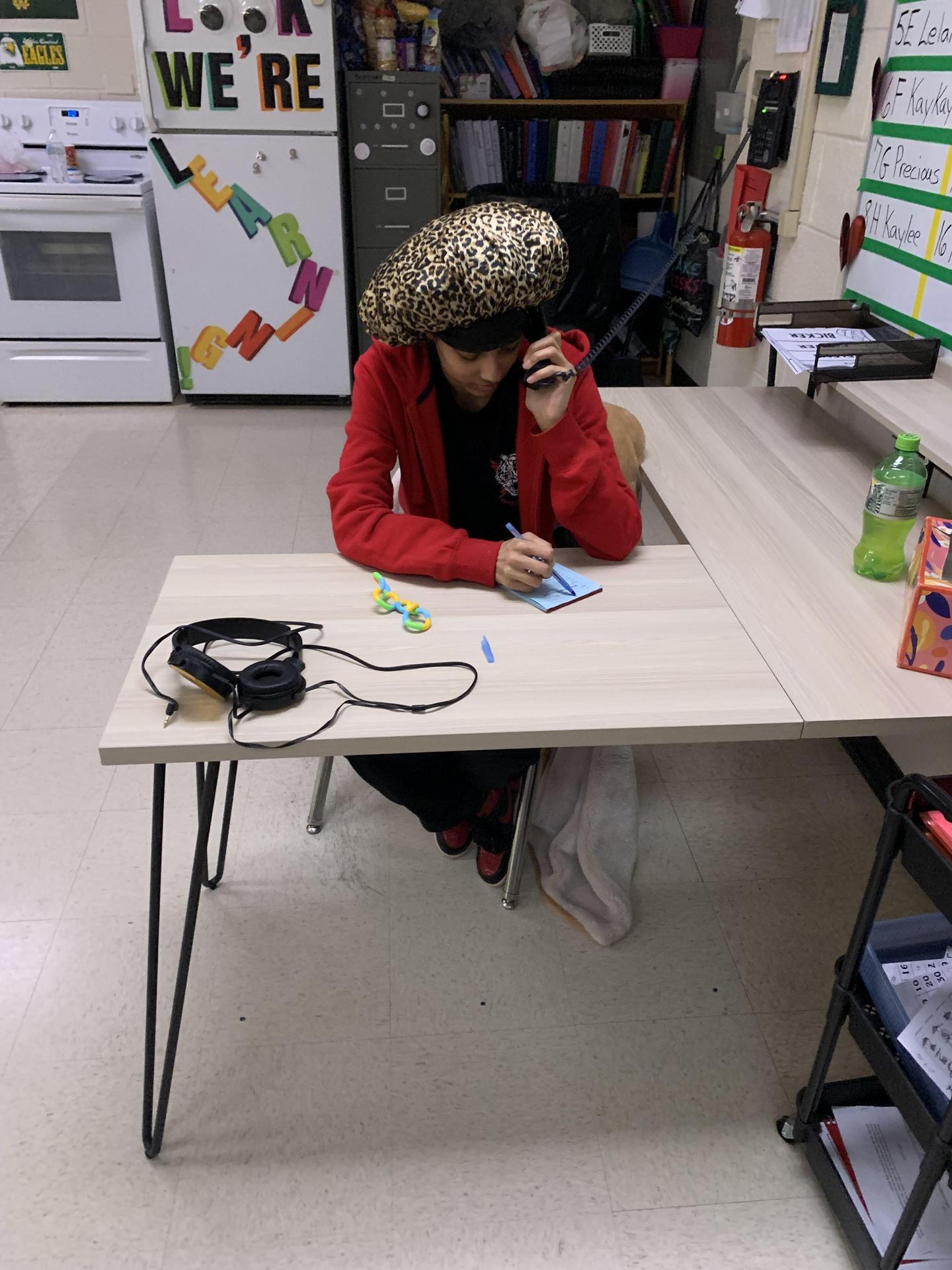 A student sitting at a table wearing a large hat while taking notes.