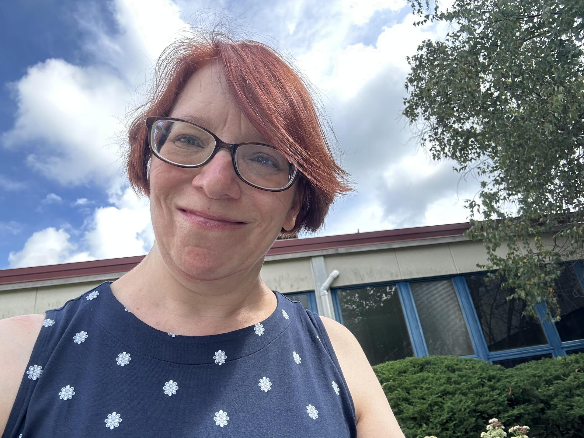 White woman with red hair and glasses in a blue shirt standing outside.